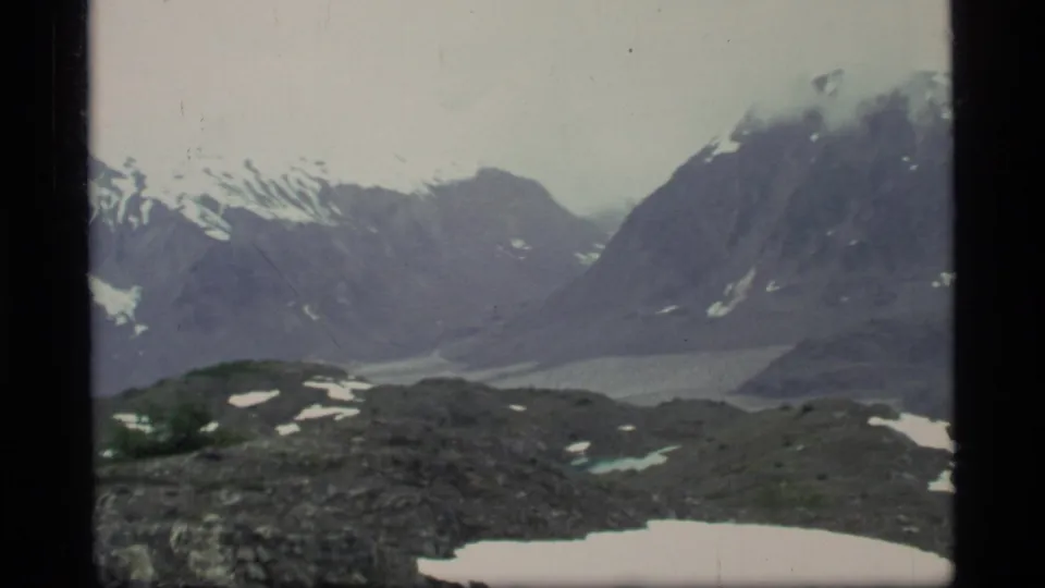 A handheld 8mm Glacier Bay landscape view shows steep mountains, snow patches, and a pale glacial valley or lake below.