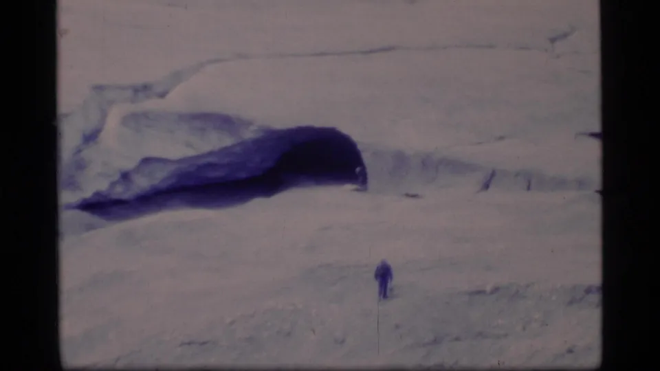A small figure walks or stands on the snow below a blue-shadowed ice cave or glacial opening in Glacier Bay.