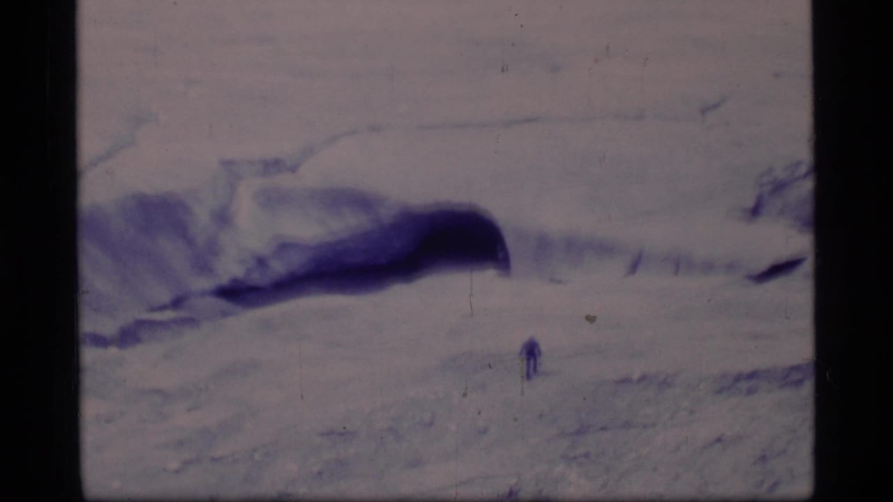 Distant person walking across a snowy glacier landscape with dark crevasses and mountains beyond.