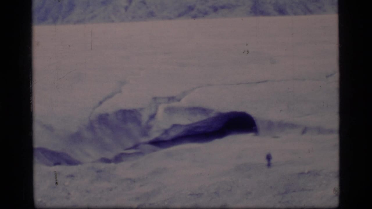 Small figure on an icy expanse beside a dark crevasse with snowy slopes in the background.