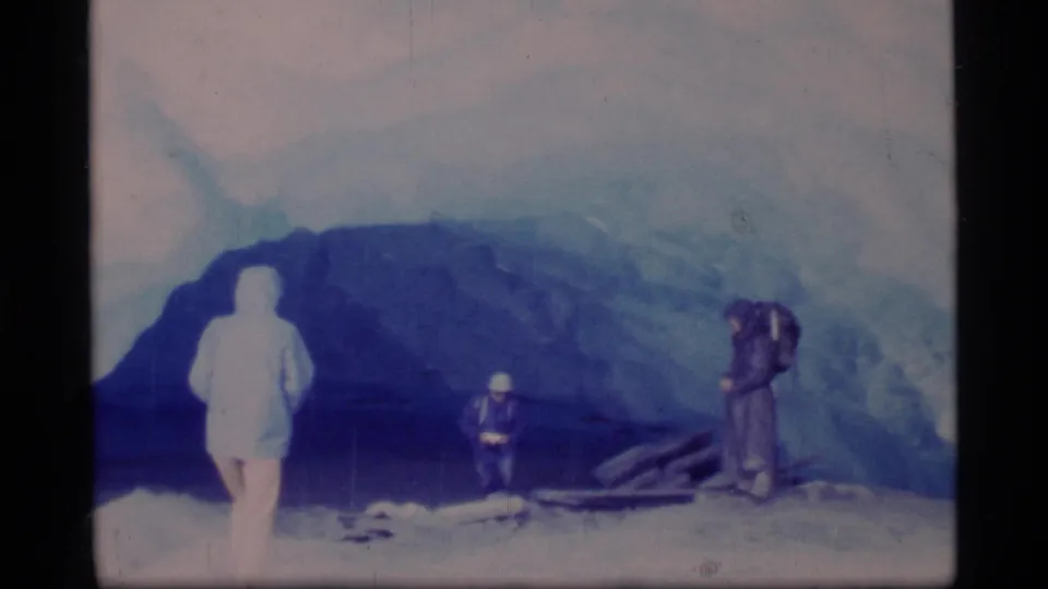 Glacier Bay 8mm frame showing three people beside a large blue ice cave, with one wearing a hooded slicker and one carrying a backpack.