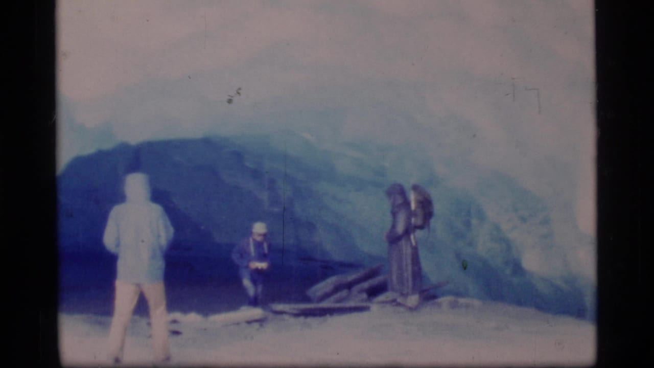 Three adults near a blue glacier, with one person appearing to film or look down at equipment.