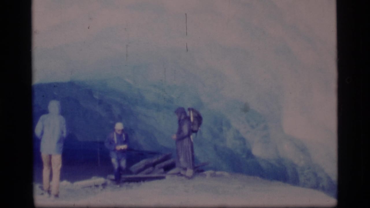Three adults stand before a glacier, including a seated figure and a person with raised arms or camera gear.