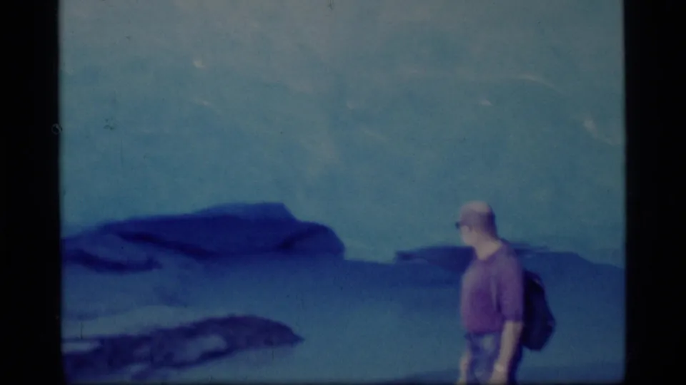 1977 Alaska home-movie frame showing a lone backpacked adult against blue glacier and mountain scenery.