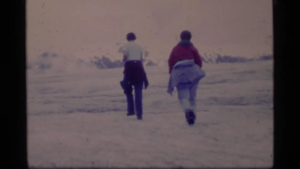Two bundled visitors walk away across Glacier Bay ice and snow in a 1977 8mm home-movie frame.