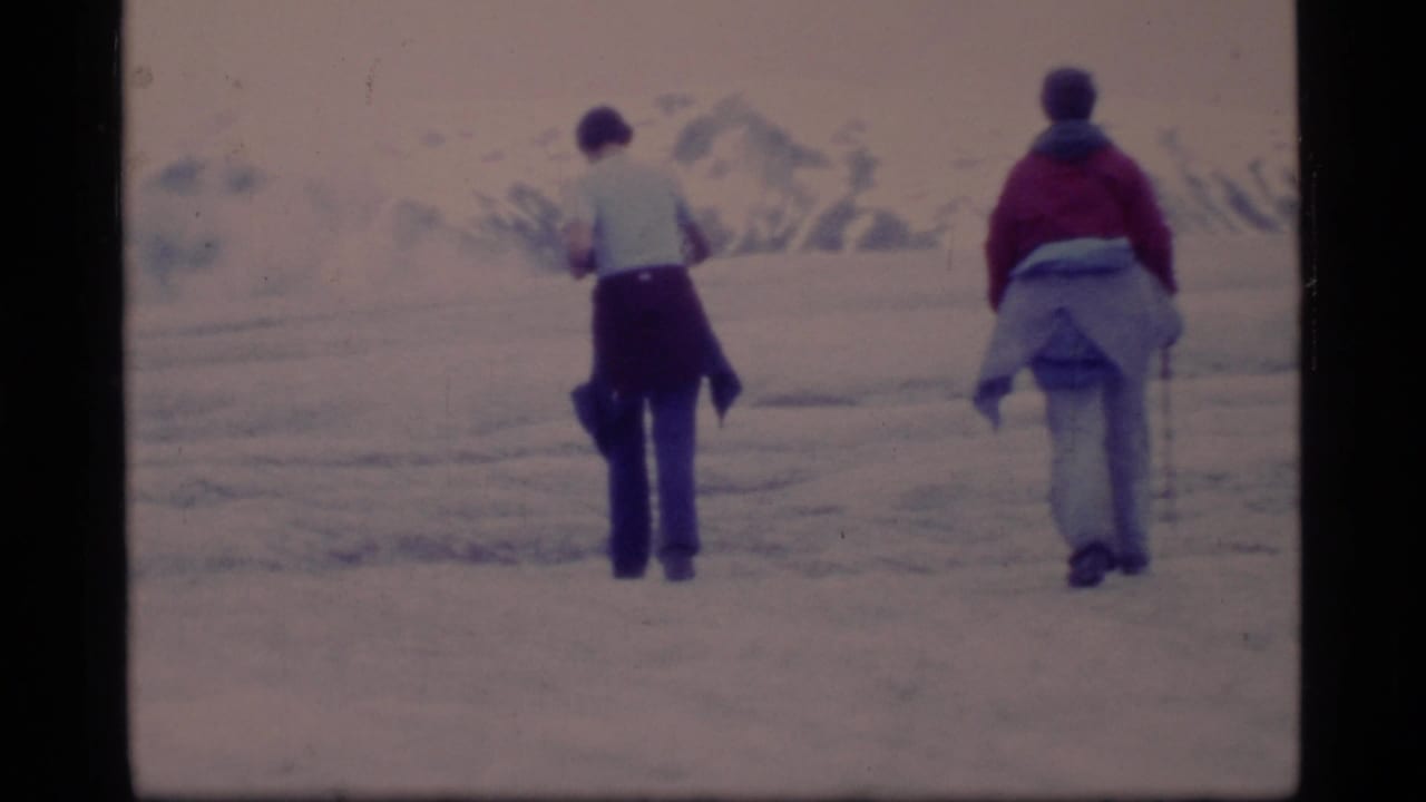 Two travelers walk away across snowy Glacier Bay terrain in 1977, with distant mountain forms and soft 8mm color fading.