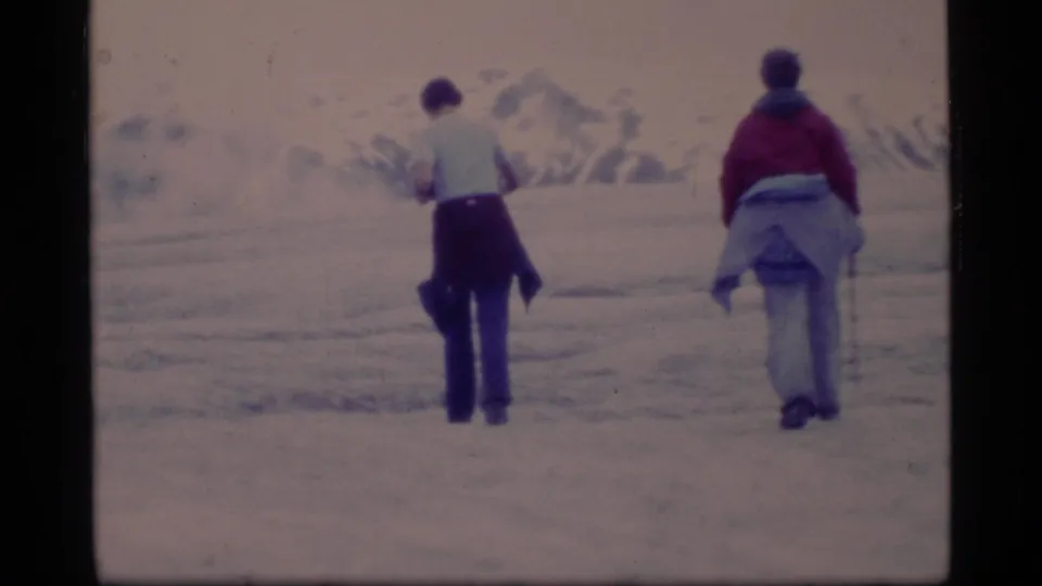 Two travelers walk away across snowy Glacier Bay terrain in 1977, with distant mountain forms and soft 8mm color fading.