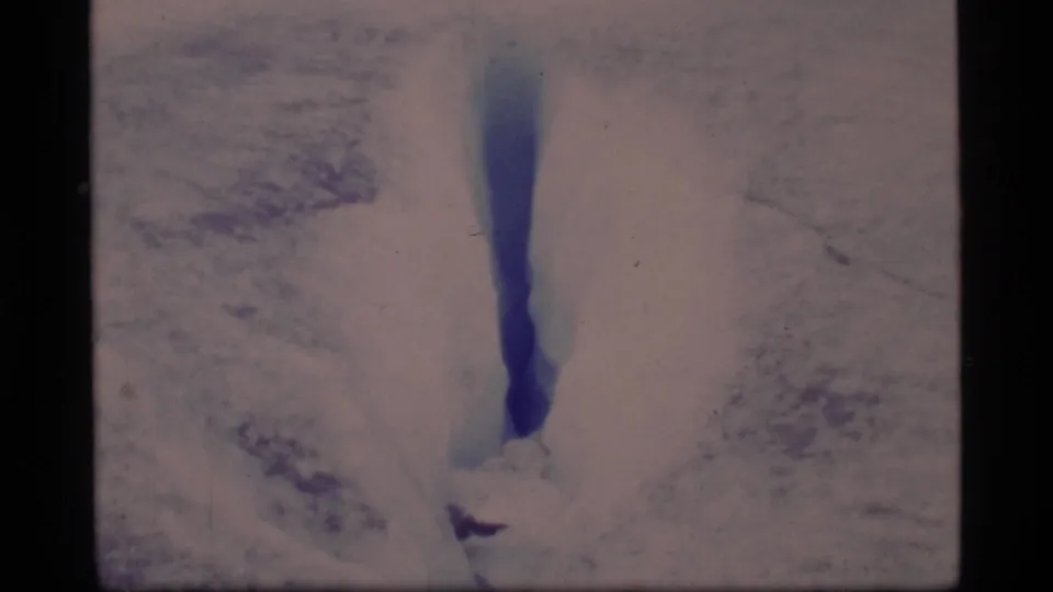The camera stays on a long, narrow crevasse in the snow, its blue center contrasting with pale glacier ice in 1977 footage.