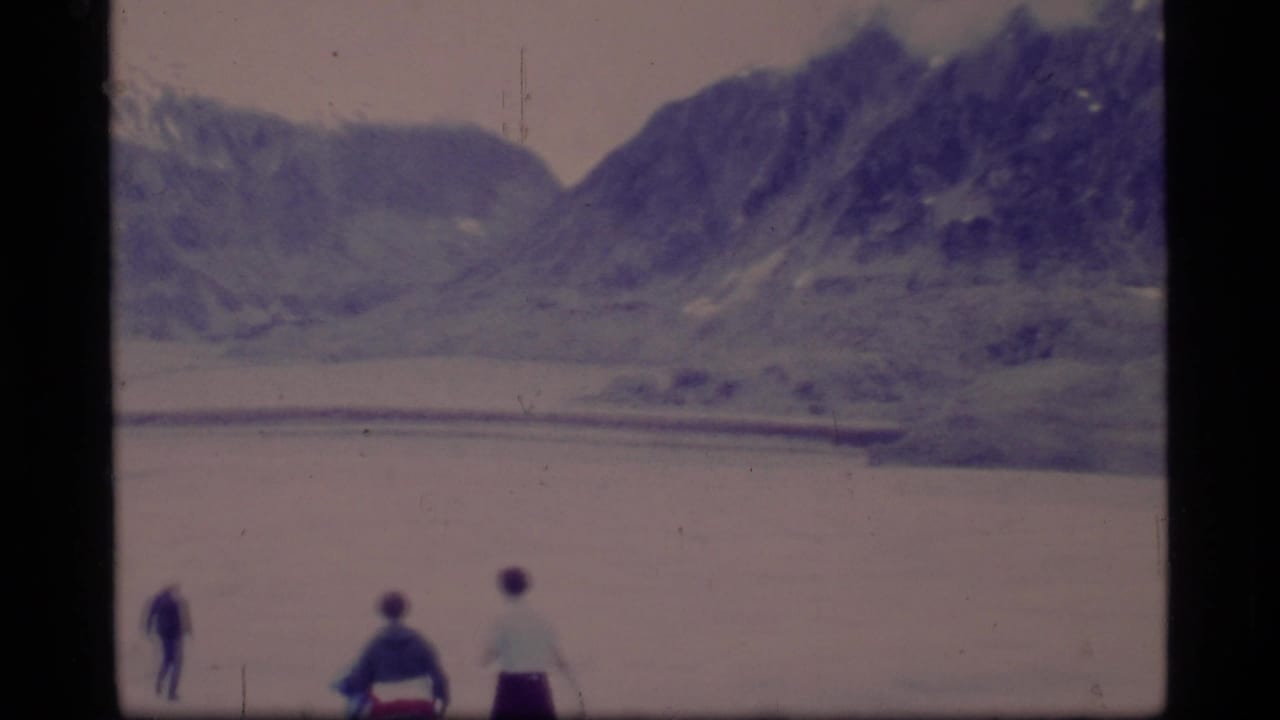 Three small people stand or walk near water below mountain ridges.