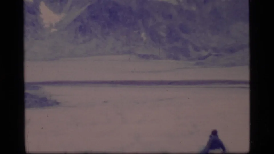 1977 8mm Glacier Bay footage showing a small distant hiker against a broad icy plain and dark mountain slope.