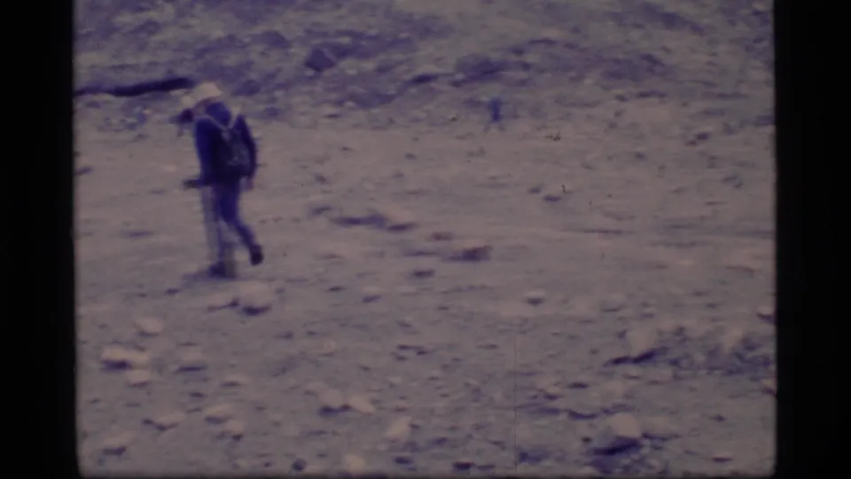 1977 8mm Glacier Bay view of a backpacked hiker crossing rocky terrain beneath a muted mountain slope.