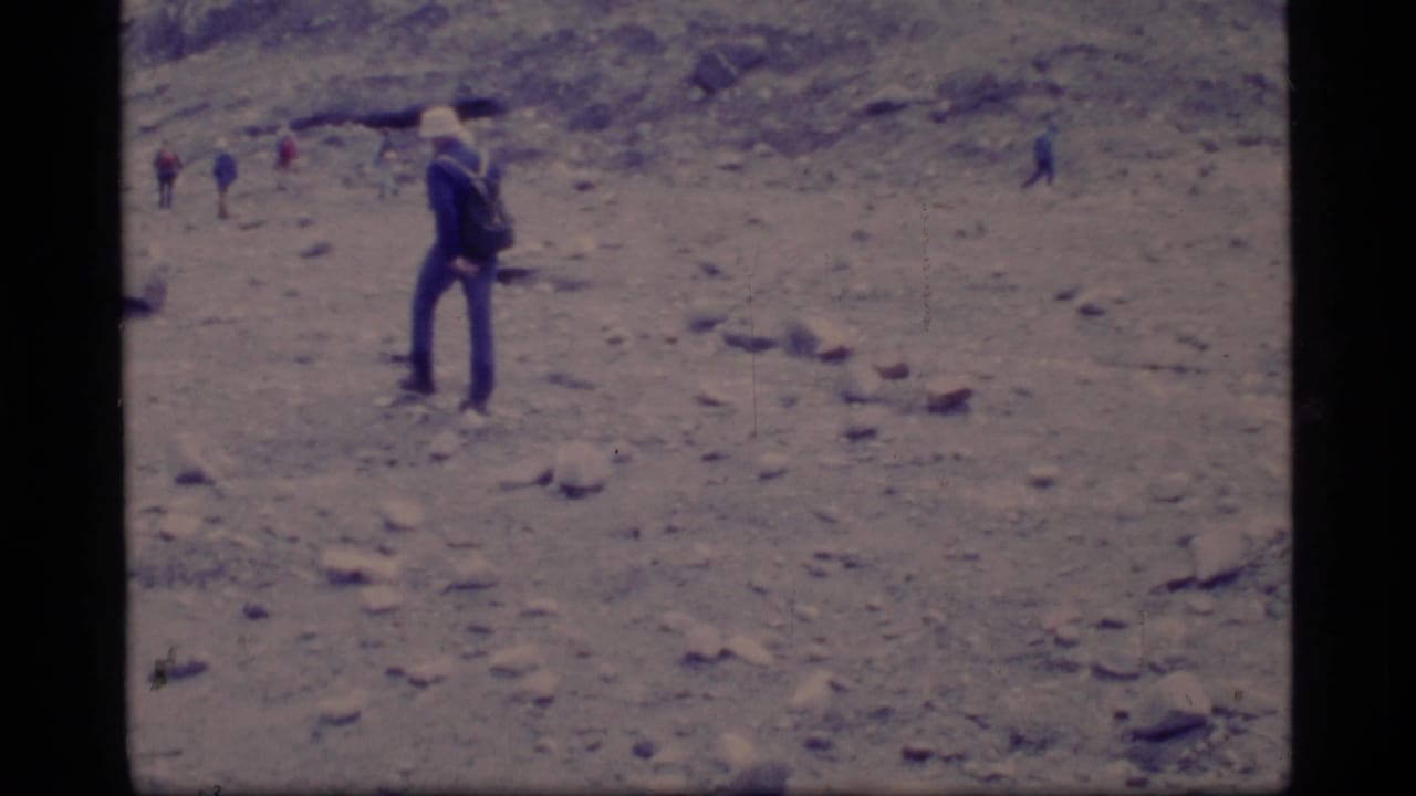 1977 Glacier Bay home-movie frame showing a cap-wearing hiker with a backpack on rocky terrain, with small figures in the background.