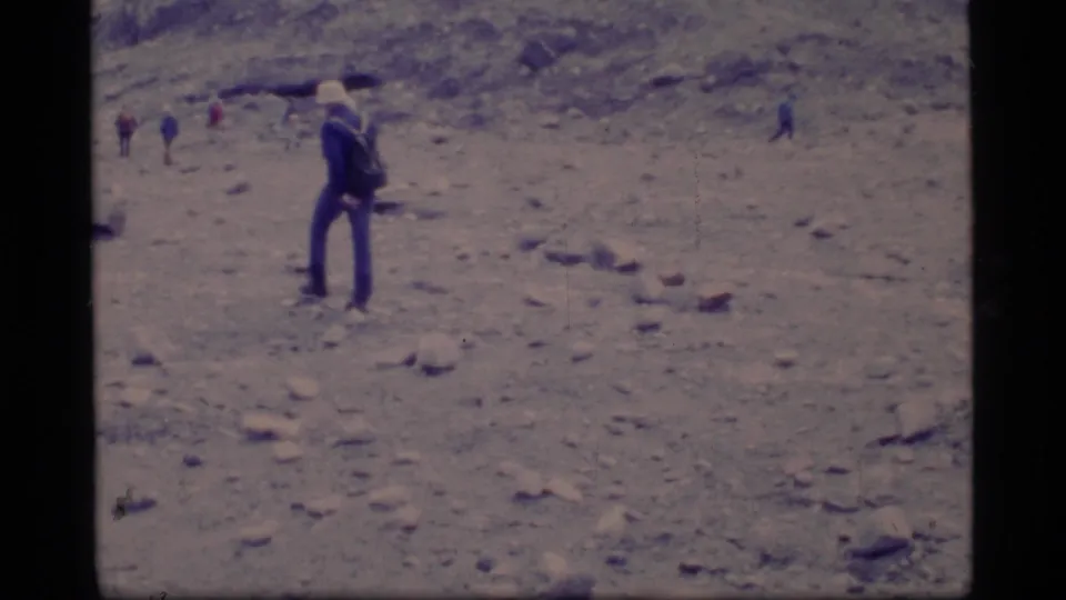 1977 Glacier Bay home-movie frame showing a cap-wearing hiker with a backpack on rocky terrain, with small figures in the background.