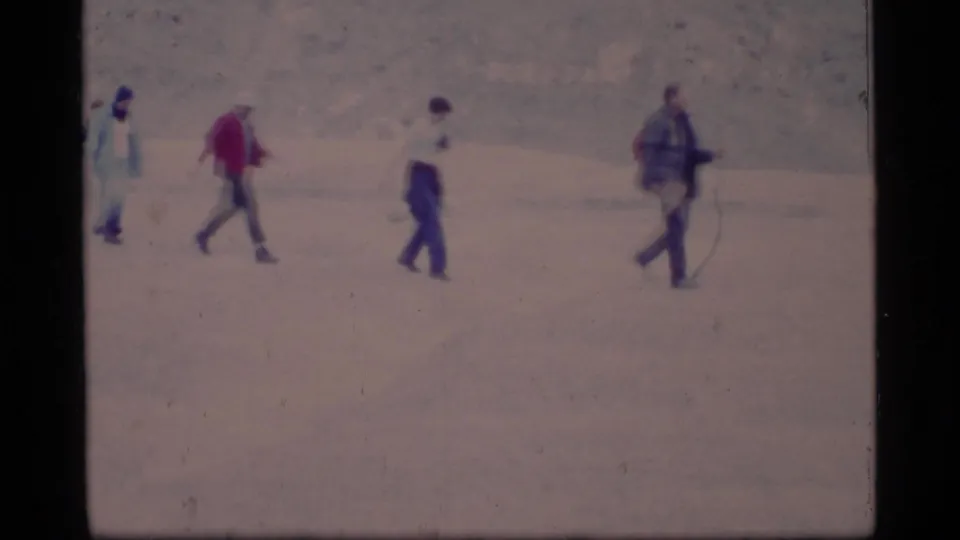 1977 Glacier Bay home-movie frame with several hikers moving across pale rocky ground, including a red jacket and a walking stick.