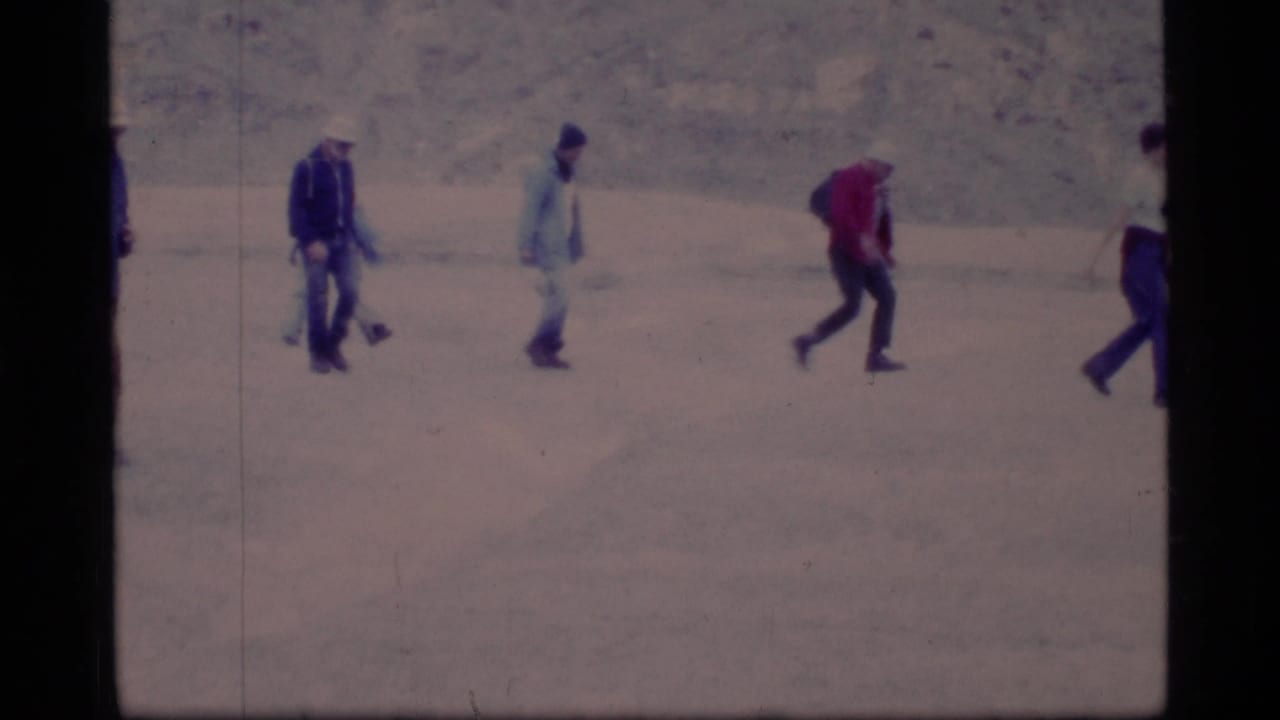 1977 Glacier Bay home-movie frame showing adults in winter clothing walking in a line across snow.