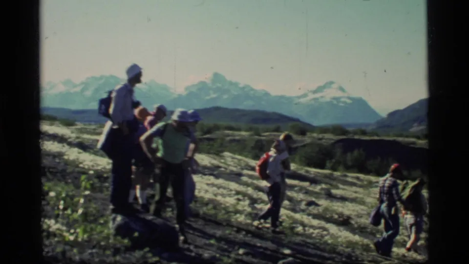 A 1977 Glacier Bay hiking group moves across uneven terrain with backpacks and snow-capped mountains behind them.