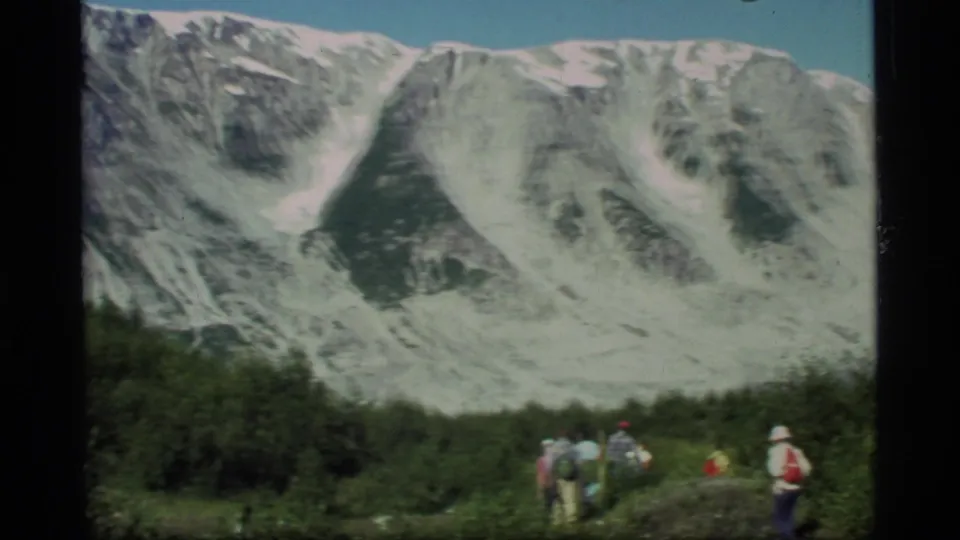 In Glacier Bay, Alaska, hikers appear small against a wide view of snow-streaked mountains and glacier-like terrain.