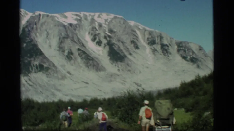 A group of hikers in 1977 home-movie footage pauses or walks through greenery beneath snowy Glacier Bay mountain scenery.