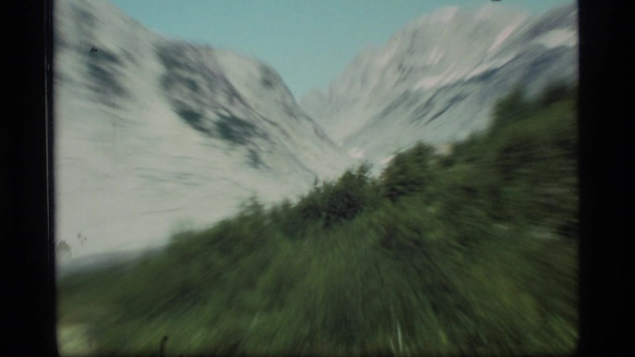 1977 8mm Glacier Bay view with snow-capped mountains and green valley brush, softened by handheld motion blur.