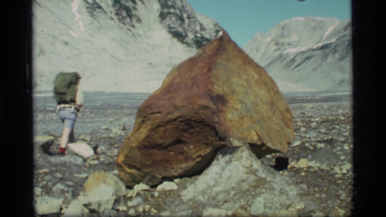 1977 Glacier Bay home-movie frame of a backpacked hiker beside a large boulder with snow-covered mountains behind.