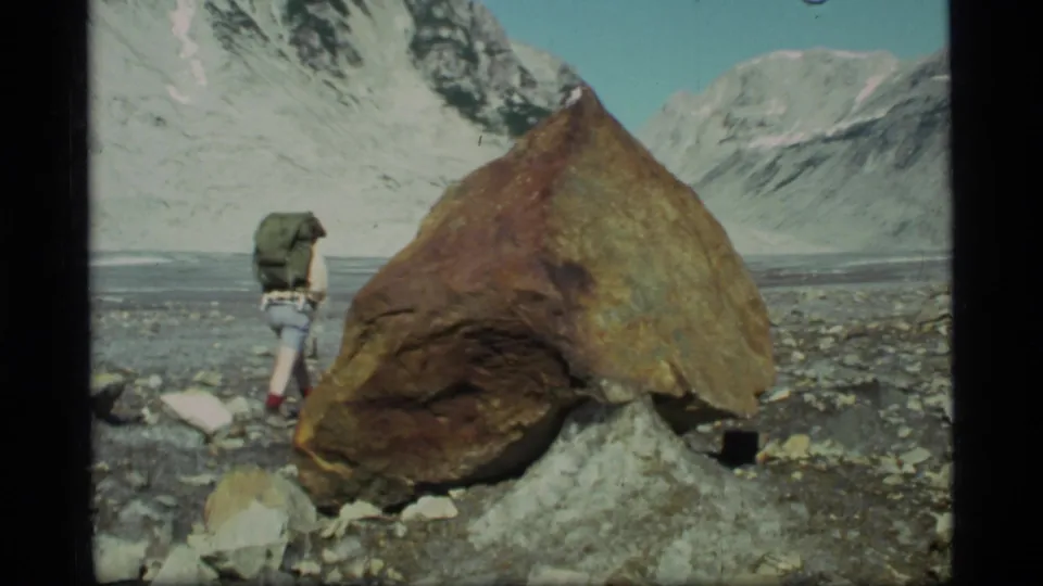 1977 Glacier Bay frame showing a backpacked hiker near a prominent rust-colored boulder and snowy mountain backdrop.