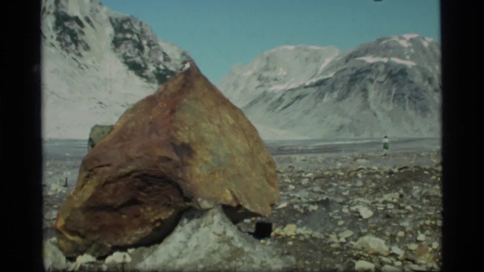 1977 Glacier Bay home-movie view with a large foreground boulder and a distant hiker in a broad rocky mountain valley.