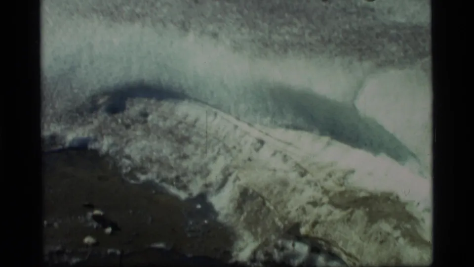 1977 Glacier Bay home-movie frame looking down at a pale glacial ice or water edge with rocky ground beneath.
