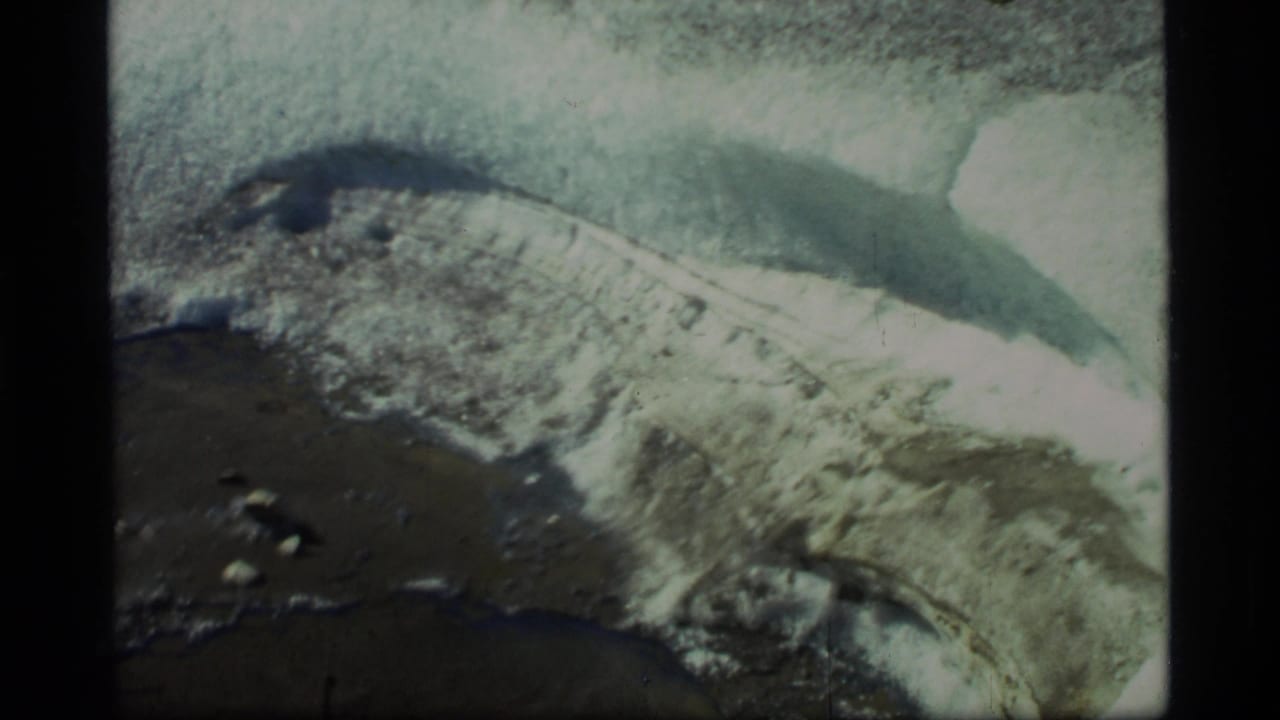 Snow and ice beside dark meltwater with rocks on wet ground.