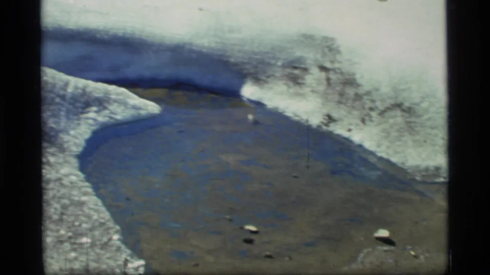 A curved meltwater pool in Glacier Bay is bordered by white snow, blue ice, and dark rocky terrain in handheld 8mm footage.