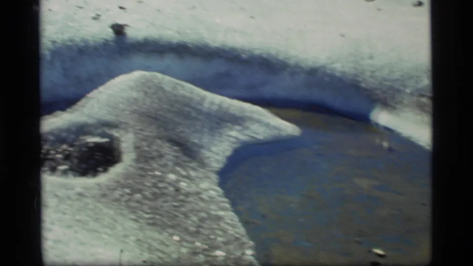 A low ridge of melting snow borders dark water and icy ground, with small stones visible in this 1977 Glacier Bay home movie frame.
