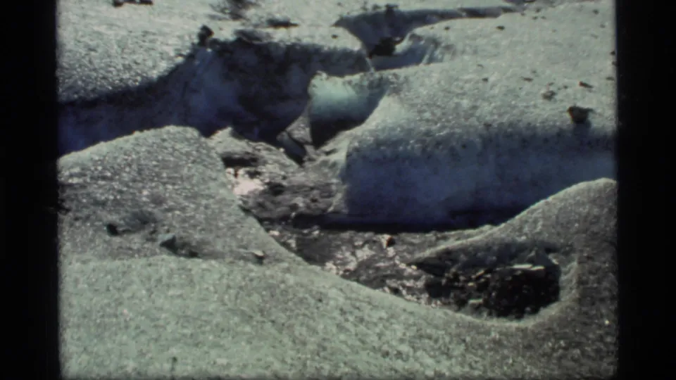 Glacier Bay home-movie frame with icy melt cuts in the foreground and distant people crossing the snowfield.