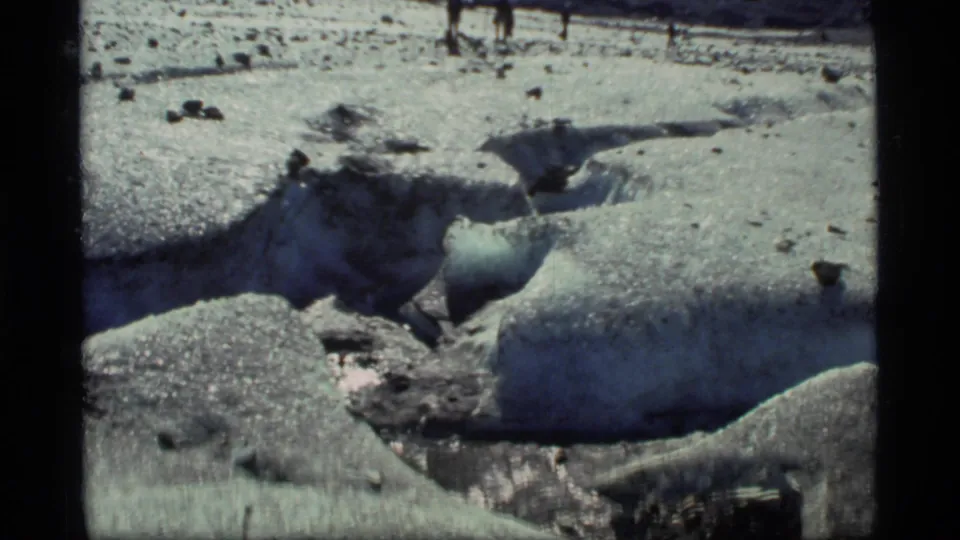 1977 Glacier Bay 8mm view of several people walking across a rough glacier surface with melt grooves and mountain backdrop.