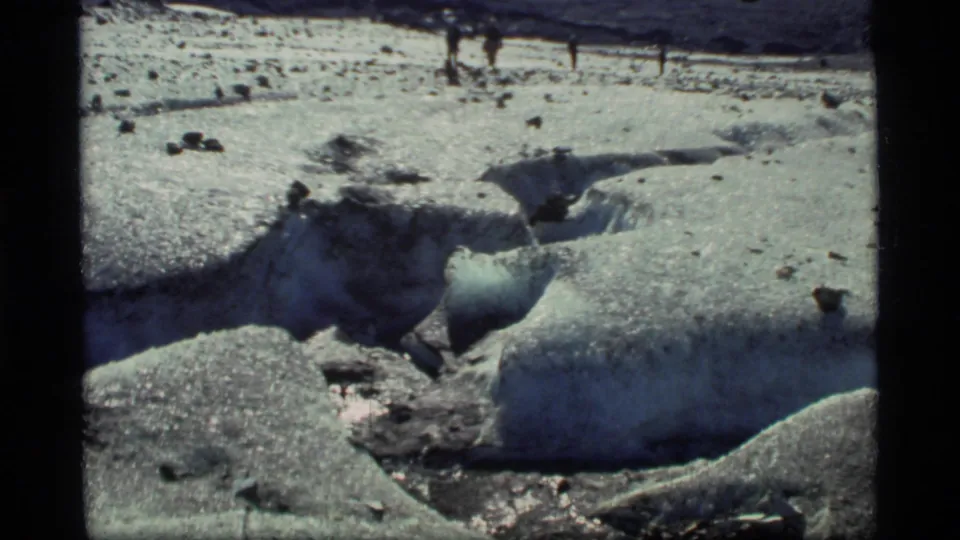 Glacier Bay frame showing a broad icy surface, dark debris, melt channels, and small walkers near the upper edge of the snowfield.