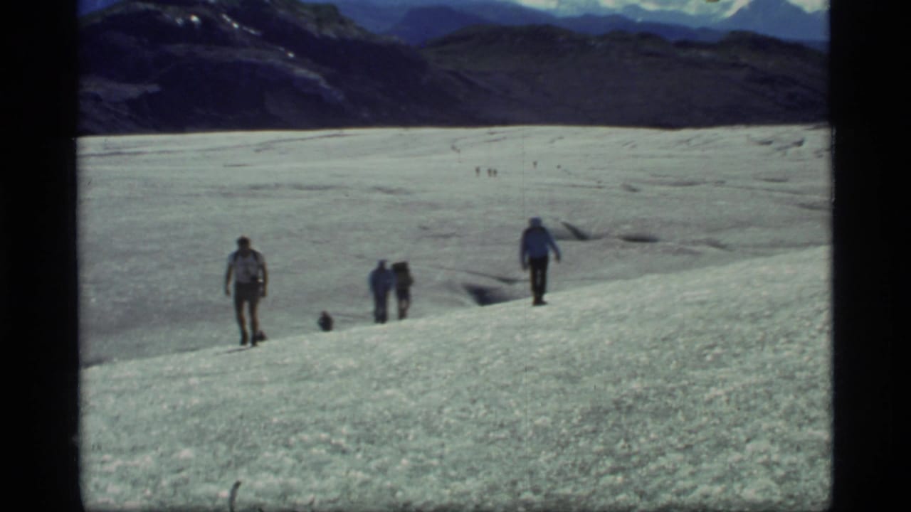 1977 Glacier Bay home-movie frame showing hikers spread across a broad glacier, with dark ridges and distant snowy peaks behind them.