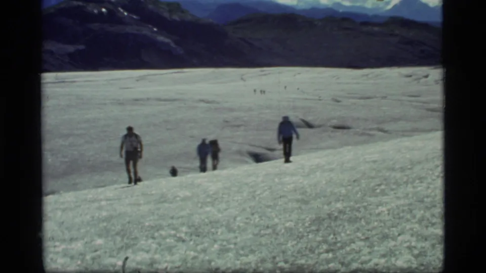 1977 Glacier Bay home-movie frame showing hikers spread across a broad glacier, with dark ridges and distant snowy peaks behind them.