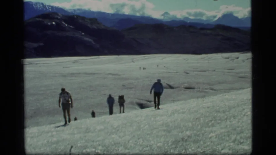 1977 Glacier Bay frame with several hikers on textured glacier ice, including one figure nearer the camera and mountains across the horizon.