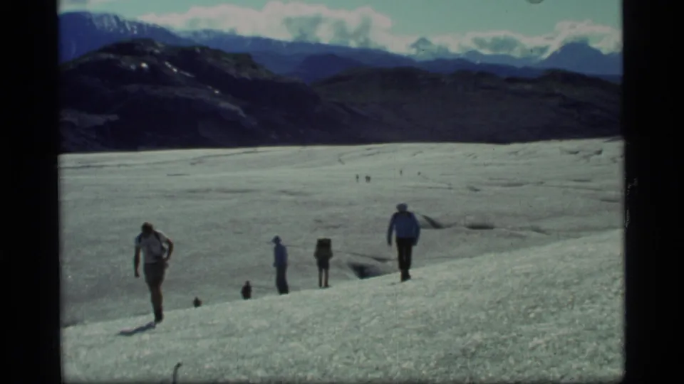 1977 Glacier Bay home-movie frame showing backpackers spaced along a glacier slope, with distant ridges, peaks, and a cloudy sky.