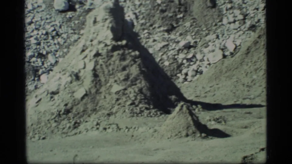 Barren Glacier Bay landscape with a pale gravel bank, scattered rocks, and strong shadow in 1977 home-movie footage.