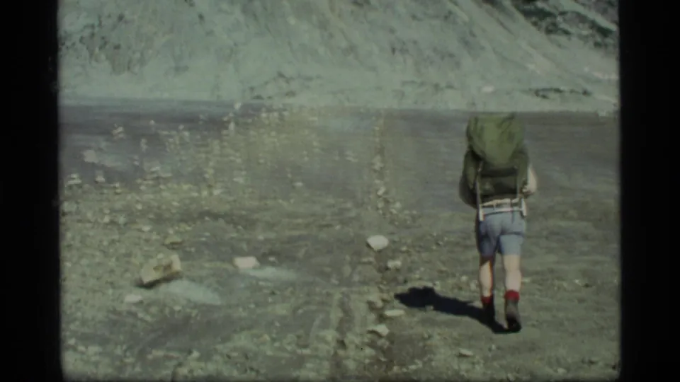 A backpacked hiker walks away across barren rocky ground in Glacier Bay, Alaska, in 1977 8mm footage.