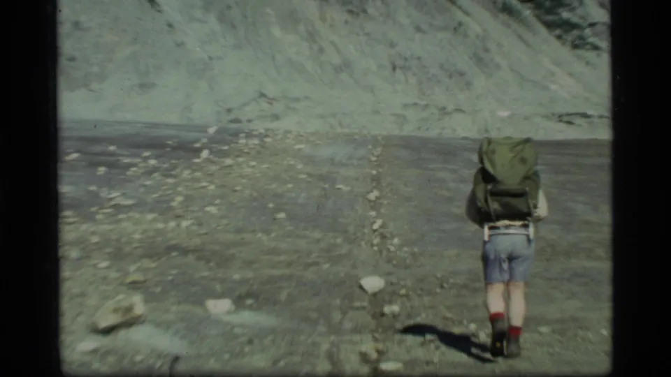 A hiker in Glacier Bay, Alaska in 1977 walks away from the camera over dry rocky terrain with a large green backpack.