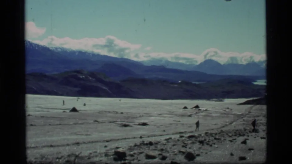 A wide 1977 Glacier Bay view shows a broad rocky plain, small distant figures, dark hills, and snow-capped mountains under blue sky.