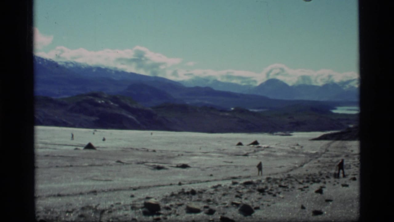 1977 8mm Glacier Bay view of distant men crossing an icy, rocky expanse below snow-capped Alaska mountains.