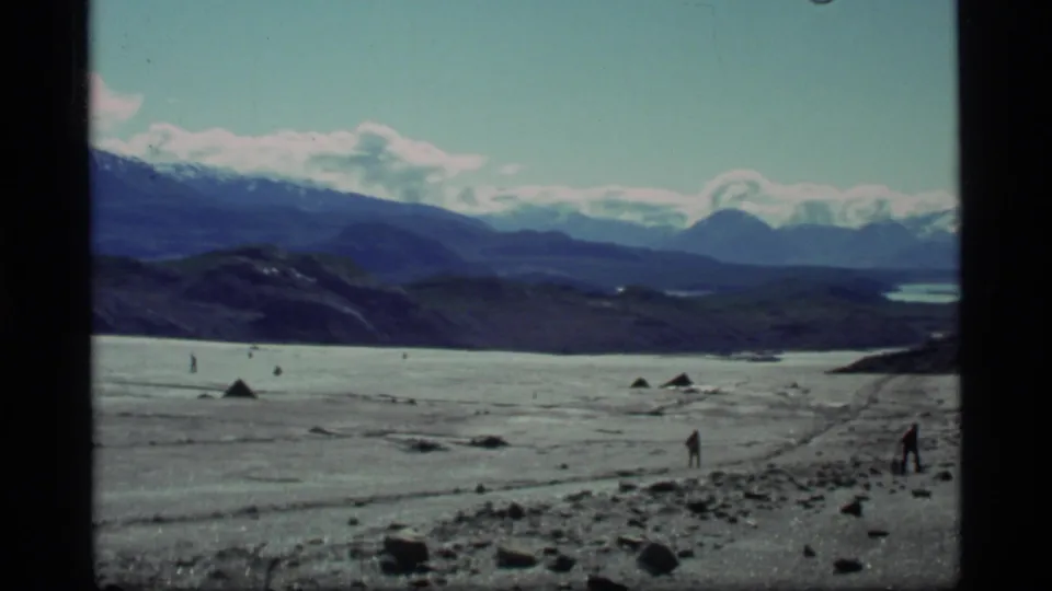 1977 8mm Glacier Bay view of distant men crossing an icy, rocky expanse below snow-capped Alaska mountains.