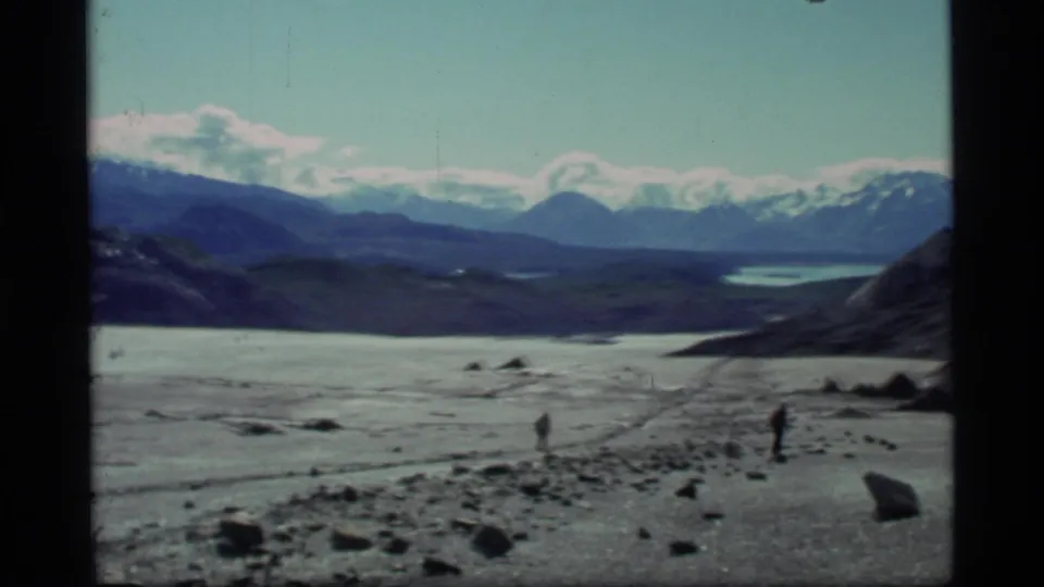 Glacier Bay home-movie frame showing two distant men on a rocky ice field with blue ridges and snow-covered peaks behind.