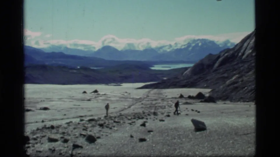 1977 Glacier Bay 8mm still of two men crossing an icy shoreline or glacier edge beside rocks and mountain slopes.