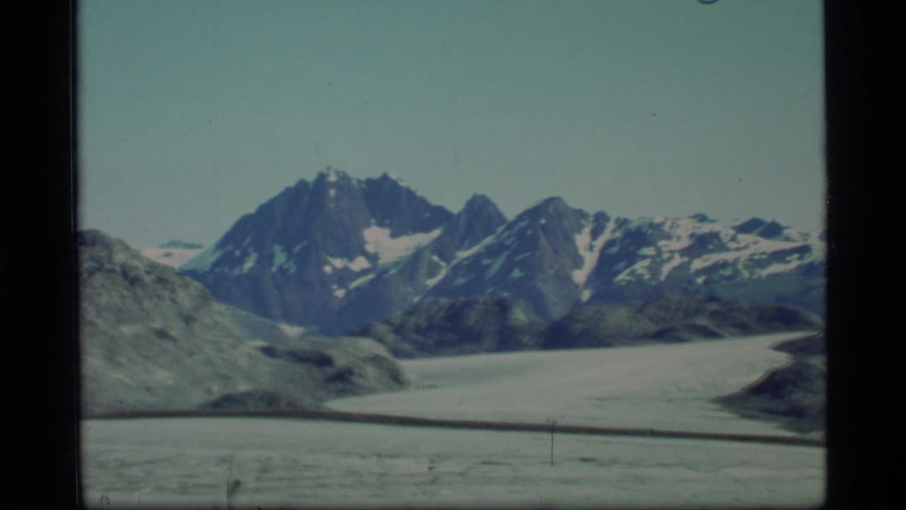 Distant snow-dusted mountain peaks rise above a broad glacier and rocky foreground.