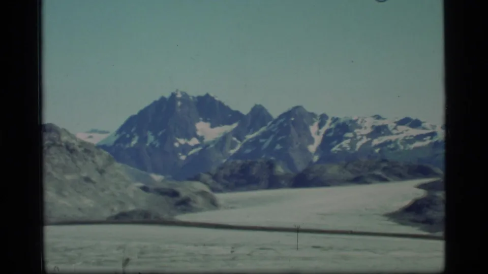 1977 8mm Glacier Bay view of rugged mountain peaks beyond a pale glacier, with rocky terrain in the foreground.