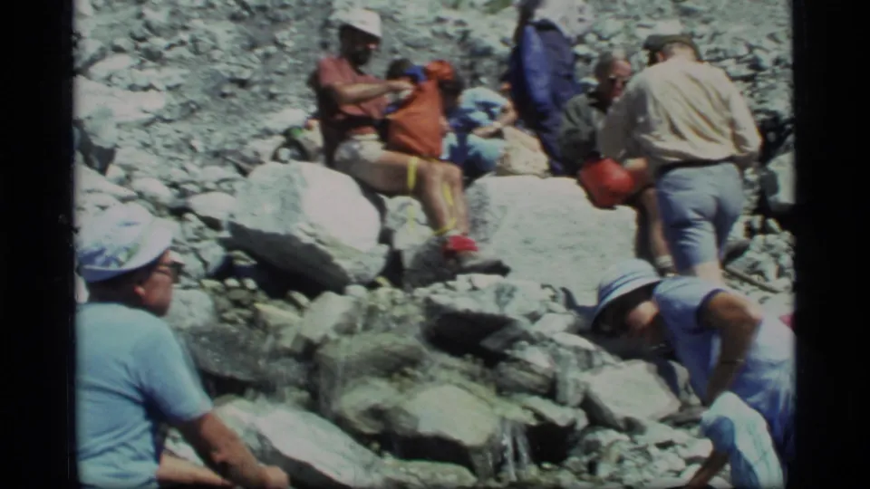 1977 Glacier Bay frame with adults in casual hiking clothes seated and standing among boulders on a rocky slope.