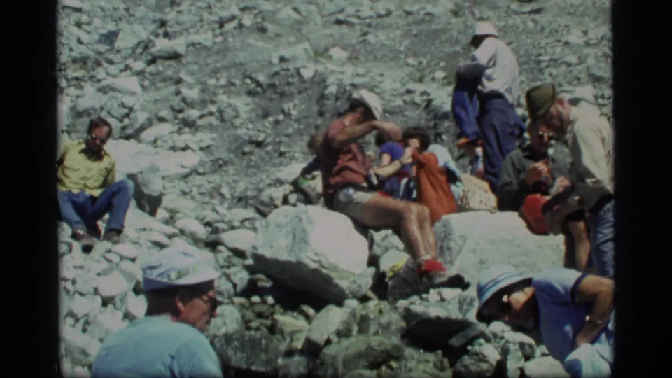 Glacier Bay 1977 home-movie frame showing a group resting on boulders, with hats and casual summer hiking clothes visible.