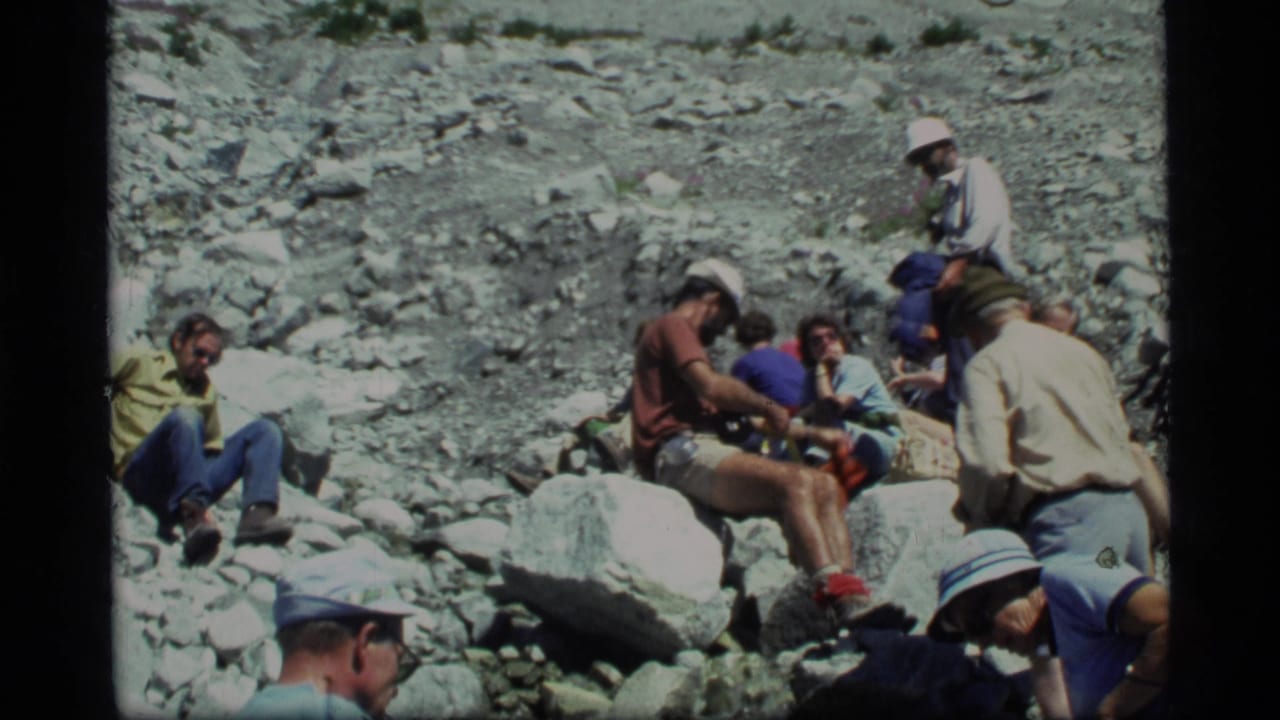 1977 Glacier Bay home-movie frame showing hikers seated and crouched among loose rocks in a mountain valley.